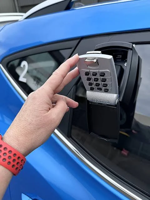 A keypad lockbox hanging from a car window, a hand opening it